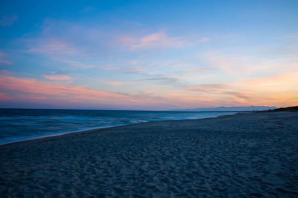 The image captures a scenic coastal sunset, showing an orange sky blending into the calm ocean. The waves roll gently onto the sandy shore, mirroring the vibrant colors of the sky.