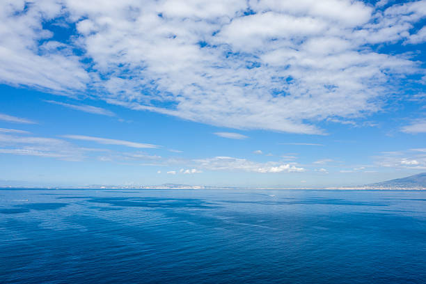 Wide view of the Bay of Naples with calm blue water stretching to the distant coastline under a sky filled with scattered clouds. The scene captures tranquil Mediterranean light and the subtle textures of open sea and horizon.