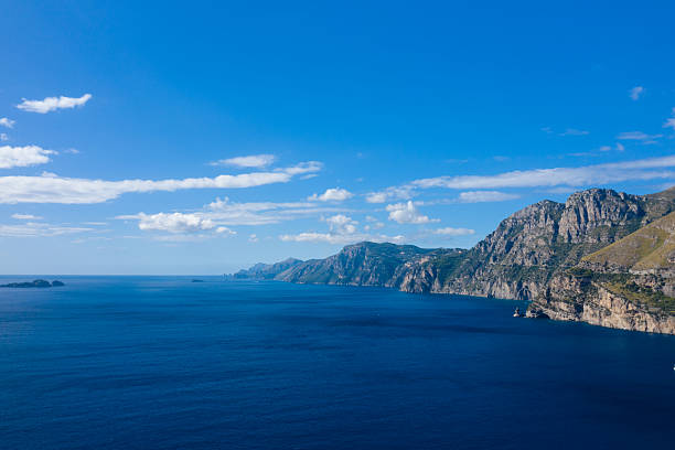 Sweeping view of rugged coastal cliffs meeting the vivid blue Mediterranean Sea under a clear sky, with scattered clouds adding depth to the horizon. Sunlight highlights the natural textures and dramatic contours of the Italian shoreline.