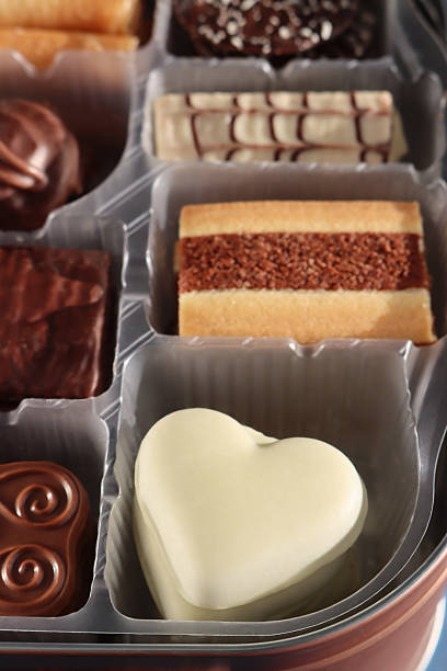Stock photo showing close-up, elevated view of an assortment of luxury collection of Belgian chocolate biscuits in a disposable, single use plastic tray inset of a biscuit tin, pictured against a mottled blue background.