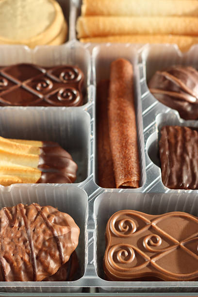 Stock photo showing close-up, elevated view of an assortment of luxury collection of Belgian chocolate biscuits in a disposable, single use plastic tray inset of a biscuit tin, pictured against a mottled blue background.