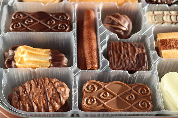 Stock photo showing close-up, elevated view of an assortment of luxury collection of Belgian chocolate biscuits in a disposable, single use plastic tray inset of a biscuit tin, pictured against a mottled blue background.