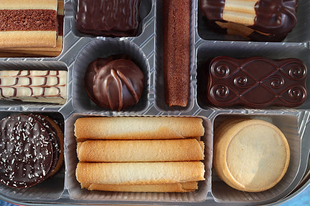 Stock photo showing close-up, elevated view of an assortment of luxury collection of Belgian chocolate biscuits in a disposable, single use plastic tray inset of a biscuit tin, pictured against a mottled blue background.