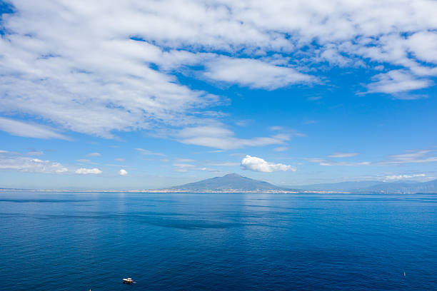 Mount Vesuvius rises in the distance across the calm blue waters of the Bay of Naples, with scattered white clouds stretching overhead. A small boat floats in the foreground, adding scale to this expansive coastal panorama.