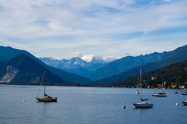 Several sailboats rest on the calm waters of Lake Maggiore in northern Italy, framed by rugged alpine mountains and a sky streaked with soft clouds. The tranquil lakeside scene features distant peaks, forested hills, and a peaceful waterfront atmosphere.