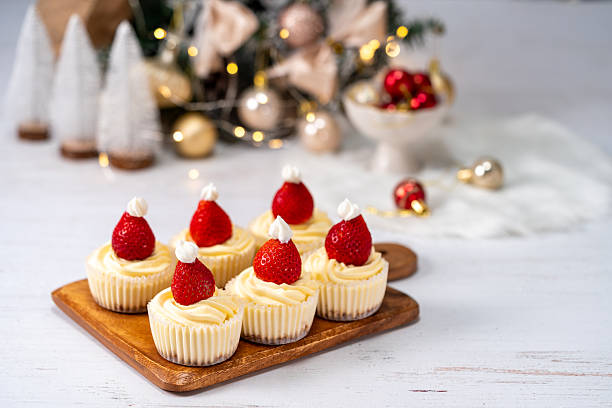 Stock photo showing close-up view of red Santa hat on top of a pile of individual mince pies, made with homemade short crust pastry, glazed with an egg wash, pictured against a background of snowy, model gingerbread houses and illuminated fir trees that form a Christmas village scene. The festive dessert tarts have been topped with pastry holly leaf detailed lid that hide the sweet mincemeat filling. Christmas festive food concept.
