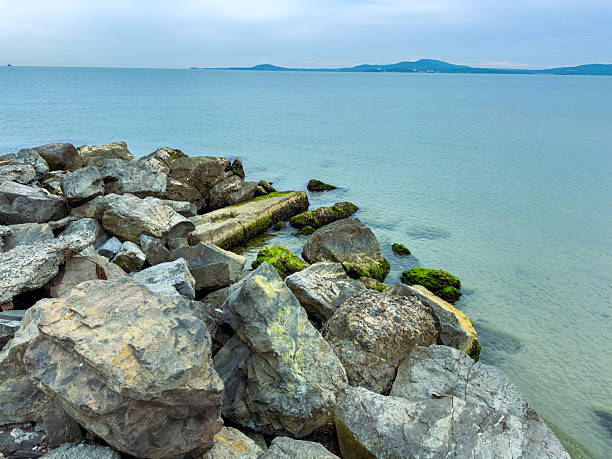Waves gently lap against a rugged coastline where large rocks create natural sculptures. Distant hills rise against a calm ocean under a soft sky, evoking peace and solitude.