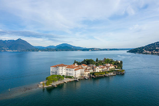 Aerial view of Isola Bella featuring its grand palace and manicured gardens surrounded by the calm blue waters of Lake Maggiore. Distant alpine mountains and lakeside villages form a scenic backdrop under a sky with soft cloud patterns in northern Italy.