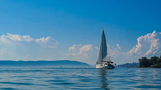 istock Yacht with a white sail on a lake. In the backgreound is a regatta and blue sky with light white clouds. Bodensee, lake Constance, Nussdorf, Germany. 2255389957