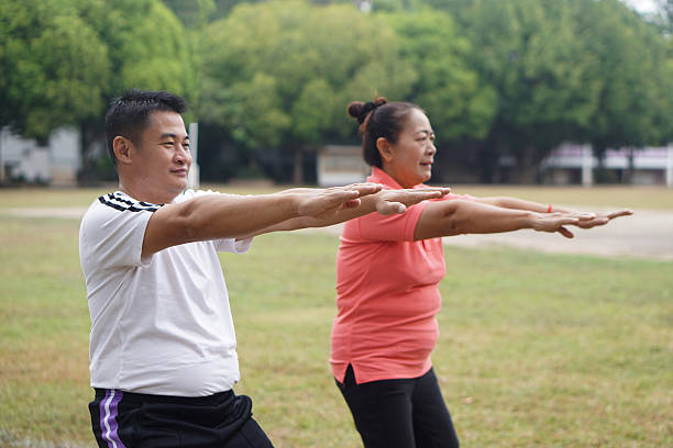 Active senior Asian couple exercising together in a public park, stretching arms forward during morning workout. Healthy aging, outdoor fitness, balance training, wellness lifestyle, retirement life