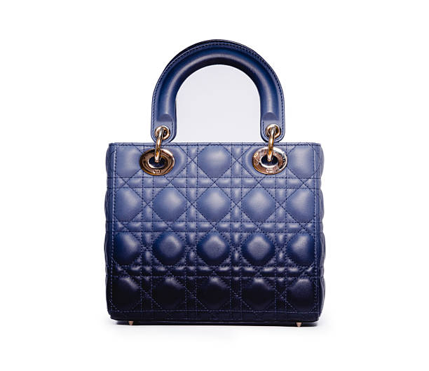 A shot of a blue leather tote bag displayed in a store window. The bag is positioned on a light-colored platform inside a display case with a dark green frame. The bag has two handles and a small pouch attached to one side. The leather appears smooth and the bag is structured. The image captures a retail display of a stylish leather bag.