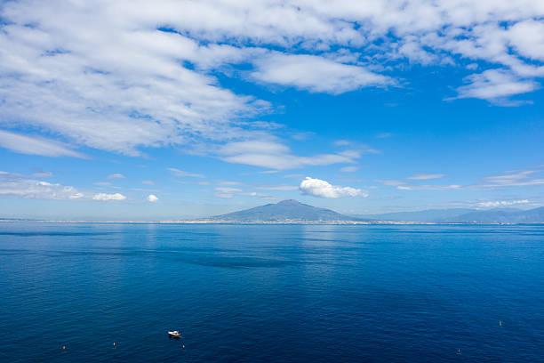 Clear blue waters of the Bay of Naples stretch toward the distant silhouette of Mount Vesuvius beneath a sky filled with scattered white clouds. A small boat floats in the foreground, adding scale to this iconic Italian coastal scene.