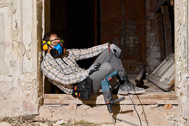 worker in respirator cutting concrete rubble inside derelict brick building, wearing earmuffs and goggles, plaid shirt and gloves,