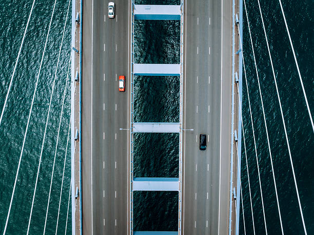 Aerial drone shot of a large suspension bridge with multiple lanes full of cars, spanning over blue-green water. Symmetrical cables frame the road deck, emphasizing engineering and travel.
