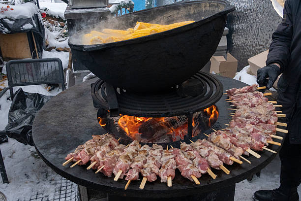 A vibrant outdoor cooking scene featuring skewers of marinated meat being grilled over a roaring open flame, surrounded by corn boiling in a large pot on a cold day.