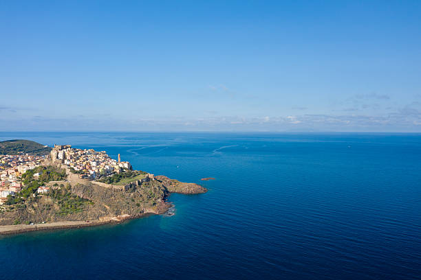 Aerial view of Castelsardo's medieval fortress and historic center rising above dramatic cliffs, surrounded by expansive azure Mediterranean waters under a bright sky.