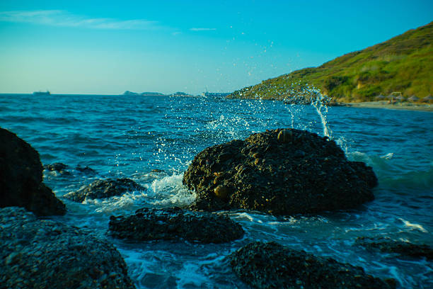 A vibrant landscape showing bright turquoise water splashing over dark shoreline rocks under a clear blue sky at Kallmi Beach.