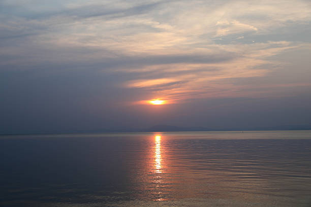 Dramatic Sunset Over Calm Sea With Soft Cloud Layers And Warm Orange Light Reflecting On Water Surface, Creating Peaceful And Atmospheric Evening Landscape With Sense Of Serenity, Nature, And Tranquil Horizon.