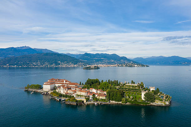 Aerial perspective of Isola Bella featuring its grand palace and ornate terraced gardens, surrounded by the deep blue waters of Lake Maggiore. Alpine mountains and lakeside towns form a scenic backdrop under a sky streaked with soft clouds in northern Italy.