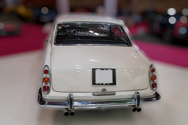 A classic white car is on display at an auto exhibition with other vintage vehicles in the background. The setting is lively as visitors admire the collection.