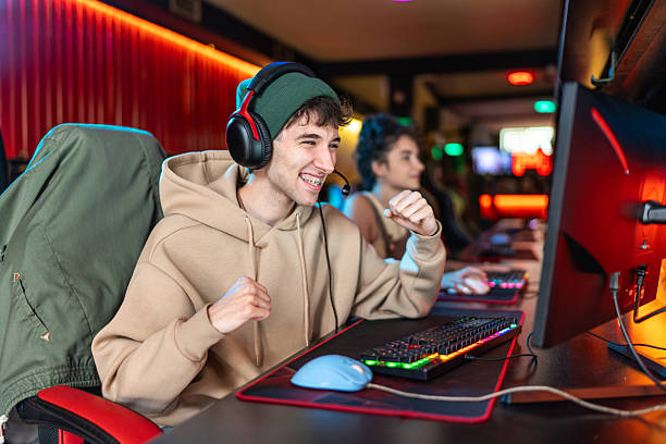 Excited young man with a headset cheering, his fists clenched in celebration, while gaming at an internet cafe with vibrant neon lighting and a female gamer in the background