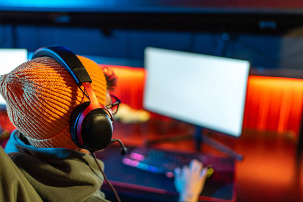 Person wearing an orange beanie and a gaming headset with a microphone playing a computer video game in a room lit with vibrant red and blue neon lights, concentrating on the monitor