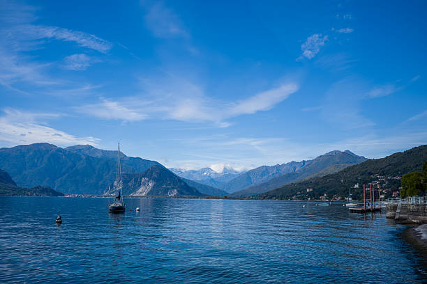 A single sailboat rests on the calm waters of Lake Maggiore in northern Italy, surrounded by forested hills and alpine peaks beneath a bright blue sky. The peaceful scene highlights the expansive lake, distant mountains, and tranquil shoreline.