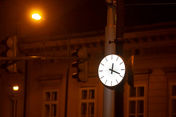 illuminated street clock on pole, glowing white face above amber traffic signals at empty intersection, historic building facade and windows