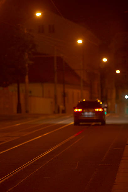 night street with car taillights glowing, empty tram tracks and wet asphalt reflecting amber lamp light, distant bokeh from streetlights,