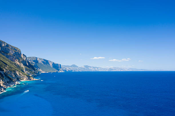 Large coastal rock formations rise above deep blue water under a bright summer sky. Soft waves and sunlight create a vivid Mediterranean scene perfect for travel and nature visuals.