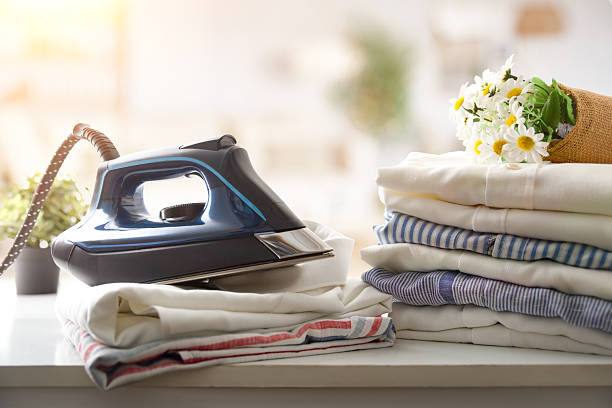 Steam iron resting on stacks of freshly ironed shirts on a white wooden cabinet with a bright room in the background. Front view.