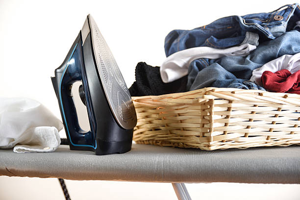 Concept detail of ironing clothes with a steam iron resting vertically on an ironing board and a basket filled with a pile of clothes. Front view.