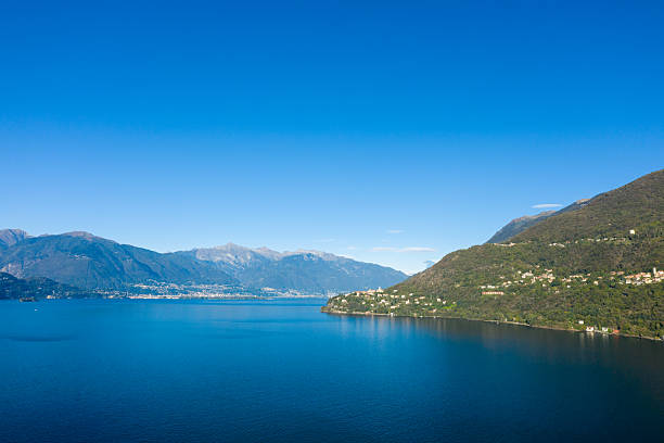 Wide view of Lake Maggiore with the hillside villages near Cannobio in northern Italy, framed by deep blue water and distant alpine peaks under a clear sky. The landscape features lush green slopes and a vibrant, tranquil atmosphere.