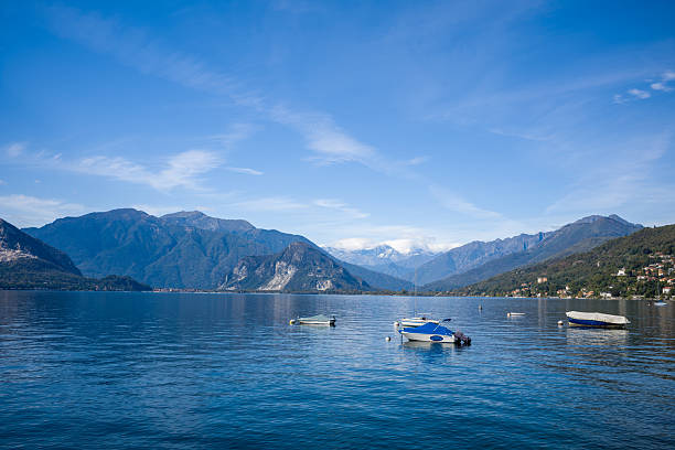 A tranquil scene of several small boats floating on the calm blue waters of Lake Maggiore in northern Italy, with rugged alpine mountains rising in the background under a clear sky. The composition highlights the expansive lake, distant peaks, and scattered lakeside houses.
