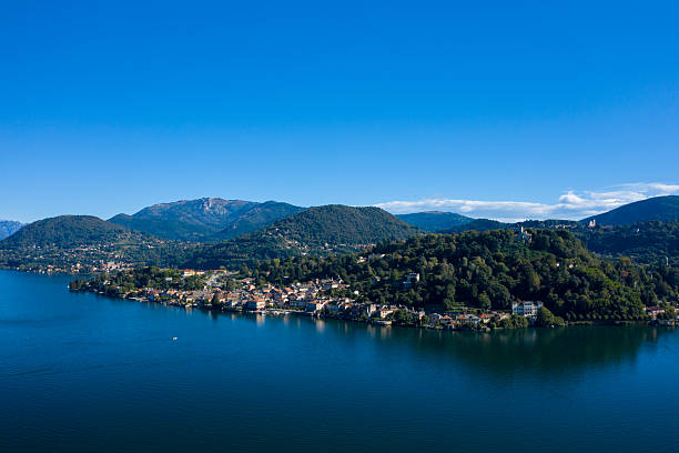 Sweeping view of a lakeside town nestled at the foot of densely forested hills along Lake Orta in northern Italy. The tranquil blue water reflects the surrounding greenery and distant mountain slopes under a vivid clear sky.