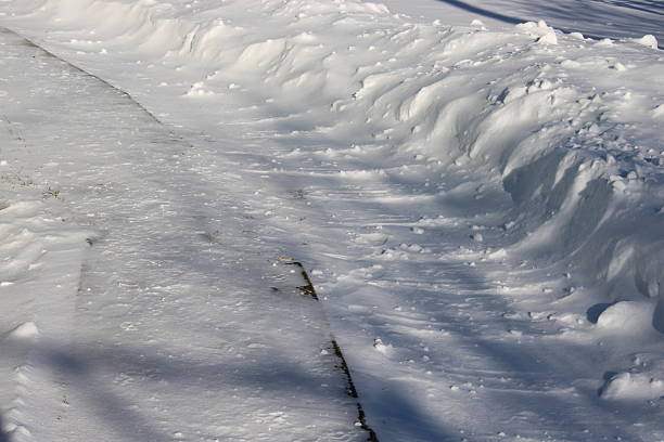 Snow is piled high on either side of a path in winter. The ground shows signs of being cleared for walking. Shadows reveal trees overhead in the bright daylight.