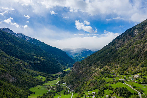 A scenic mountain road curves through a lush green valley surrounded by forested slopes and distant snow-capped peaks in Val d'Aoste, Italy. The landscape features vibrant meadows, dramatic elevation, and a mix of sunlight and clouds under a bright alpine sky.