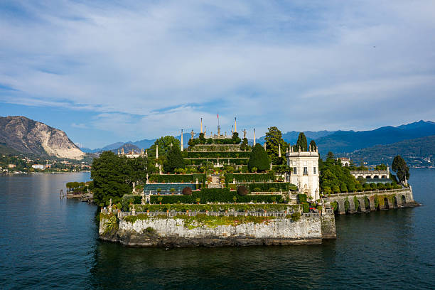 Baroque terraced gardens and a grand palace rise above the water on Isola Bella, surrounded by the blue expanse of Lake Maggiore. The island is framed by lush greenery and distant alpine mountains in northern Italy.