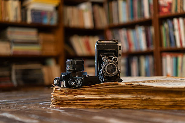 Vintage folding camera and modern film camera placed on a stack of old books on a wooden table inside a library.