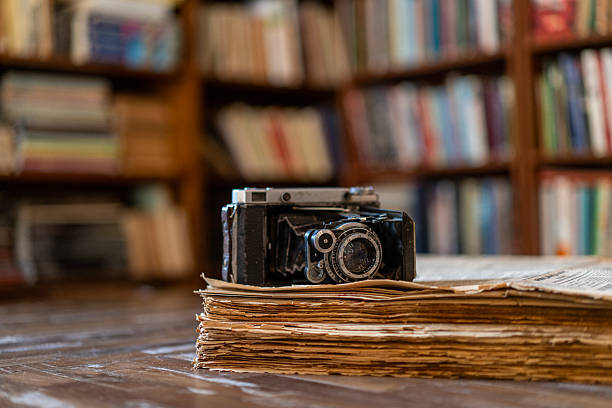 Vintage folding camera and modern film camera placed on a stack of old books on a wooden table inside a library.
