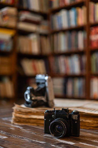 Vintage folding camera and modern film camera placed on a stack of old books on a wooden table inside a library.