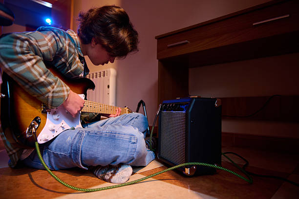 A young guitarist sits on the floor, strumming an electric guitar beside a compact practice amp in a cozy home setting, capturing focus and creativity.