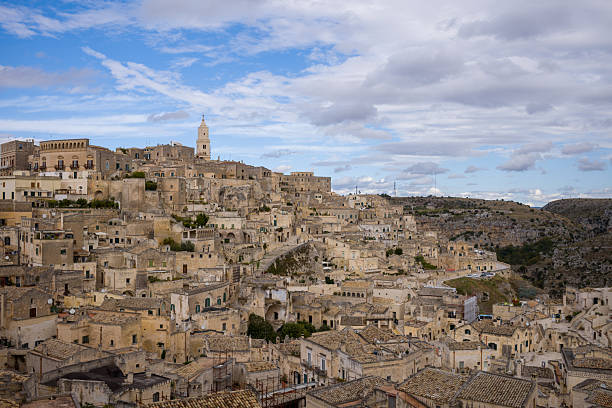Clustered stone houses and historic buildings cascade down the rugged hillside of Matera, Italy, beneath a dramatic cloudy sky. The textured architecture and earthy tones create a timeless Mediterranean landscape.
