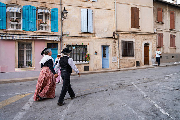 Arles, France - July 1, 2024: A senior couple walk hand in hand, the women dressed in long period gowns with lace shawls and Arlesian hats walk along the road around the Roman Arena at the end of the Costume de Fete,  Europe