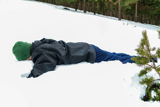 A cute laughing child dressed in a blue winter jacket, brown snow pants, and a striped knit hat with a pom-pom lies joyfully in the snow. Wearing a matching scarf and gloves, the boy smiles brightly, surrounded by a blanket of white snow. Shot with Nikon D800.