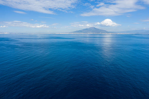 Mount Vesuvius rises in the distance beyond the expansive blue waters of the Bay of Naples, with scattered white clouds reflecting on the calm sea. The scene is defined by clear daylight, open horizon, and subtle gradients of Mediterranean color.
