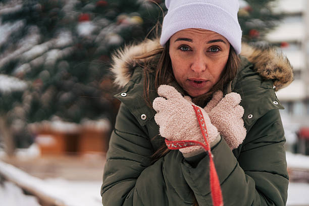 Woman feeling cold and uncomfortable, wrapping her arms around herself while standing outdoors in snowy winter conditions, wearing a warm coat, beanie, and fuzzy mittens