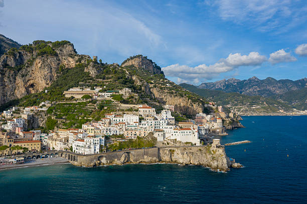 A wide view captures the sunlit whitewashed buildings of Amalfi, Italy, cascading down steep cliffs toward the deep blue Mediterranean Sea. The dramatic shoreline features historic stone arches, lush greenery, and rugged mountains under a clear sky.