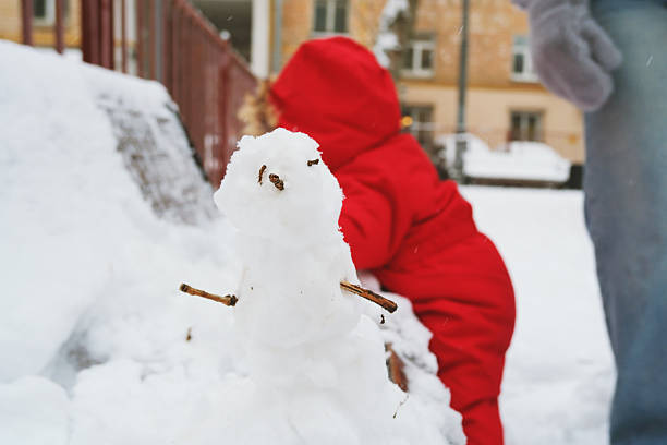 Child in a red coat interacts joyfully with snow, creating a small snowman in the playground. The scene captures playful winter fun and creativity in a snowy setting.
