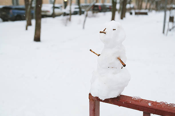 Simple snowman with stick arms and a smiling face stands near a snowy park bench with blurred winter city lights background, winter leisure